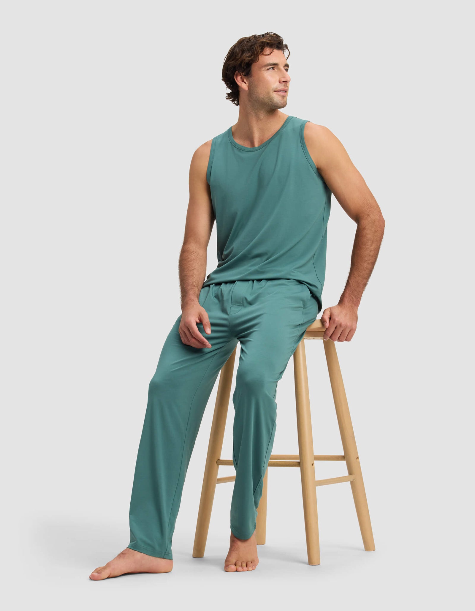 A man in Cozy Earth's Men's First Layer Pajama Pant, paired with a teal sleeveless top, sits barefoot on a wooden stool, smiling and looking left against a plain light background.