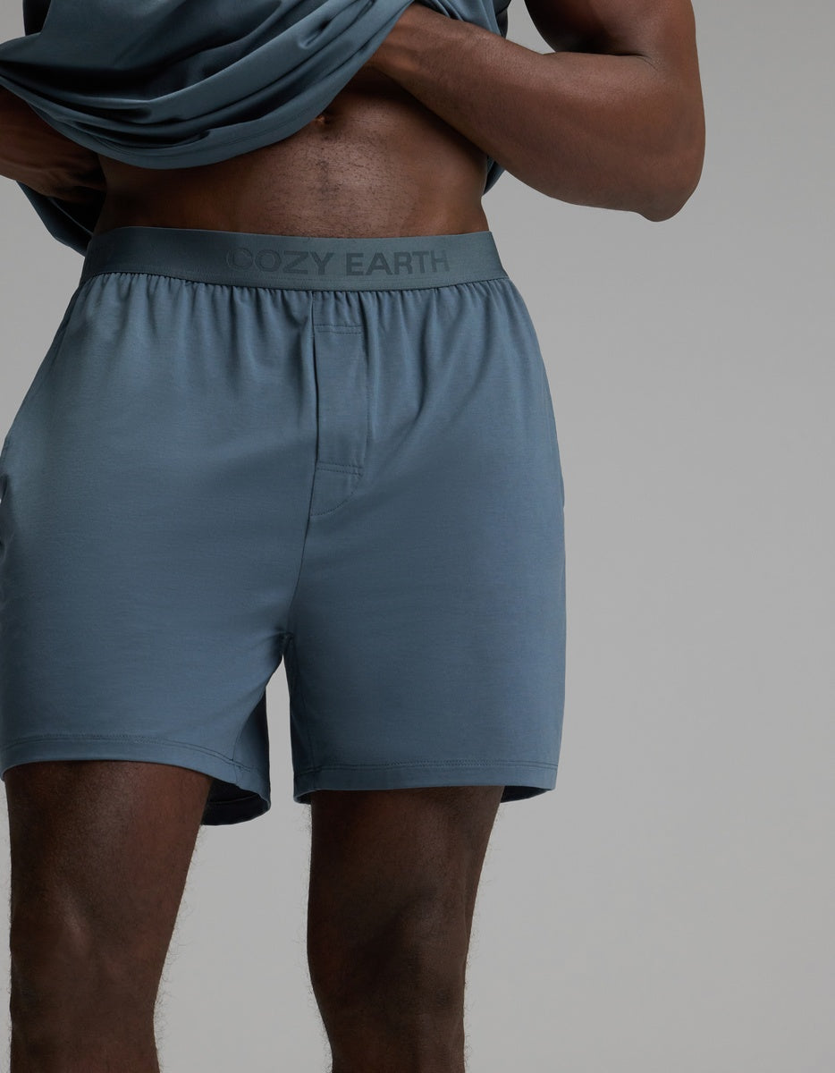 A person wears Cozy Earth’s Men’s First Layer Pajama Short in Dawn, featuring “Cozy Earth” on the waistband, lifting their shirt to reveal their torso and legs against a plain background.|Color:Dawn
