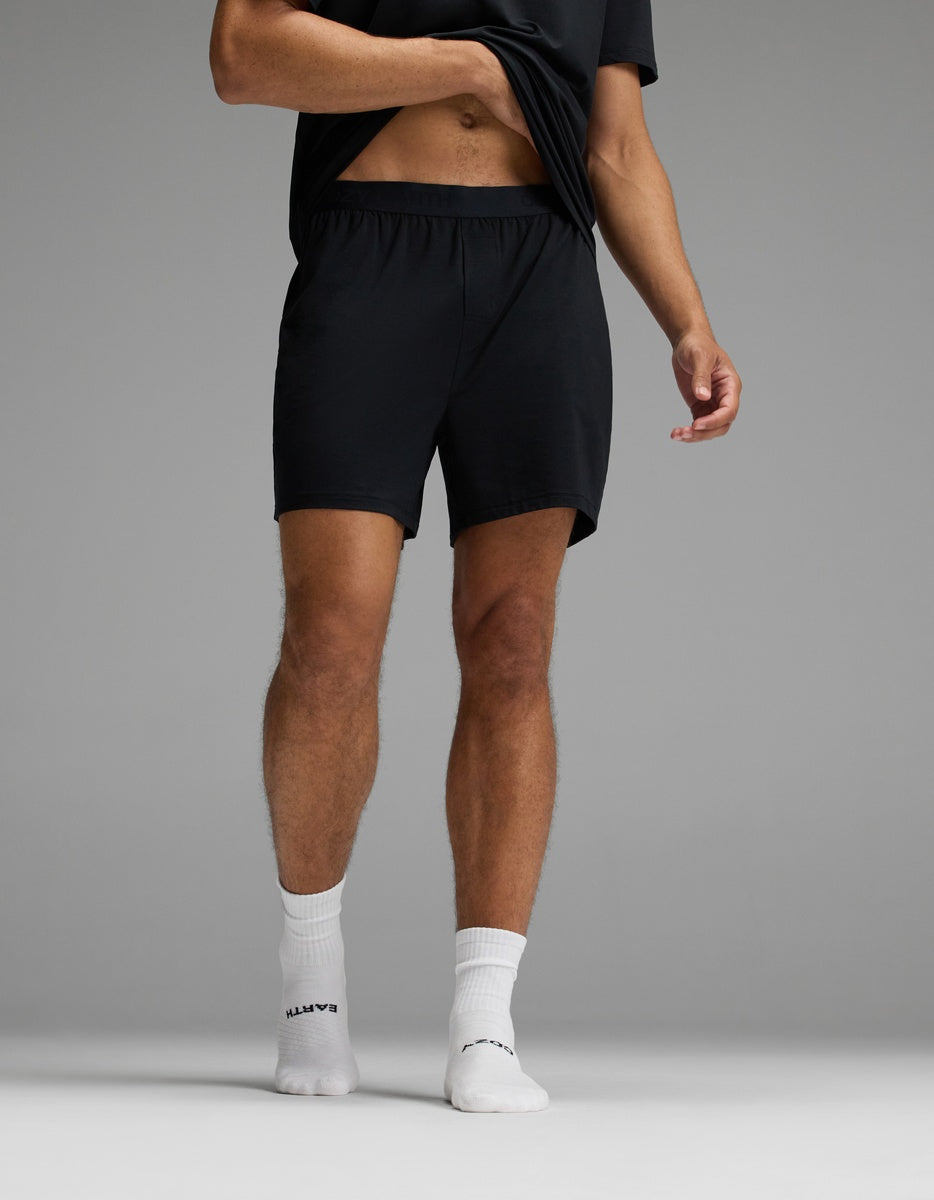 A person is standing on one foot against a plain gray background, wearing Cozy Earth Men’s First Layer Pajama Shorts paired with a black t-shirt and white socks, lifting their shirt slightly.|Color:Jet Black