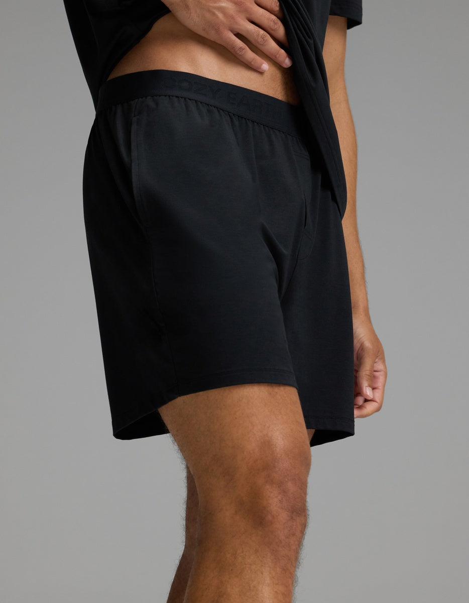 A person wearing Cozy Earth’s Men’s First Layer Pajama Short in Jet Black, paired with a matching black shirt, stands against a plain gray background with one hand resting on their waist.|Color:Jet Black