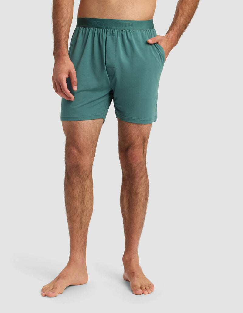 A man stands barefoot in Cozy Earth Men's First Layer Pajama Shorts with an elastic waistband, one hand in his pocket, against a plain light gray background.