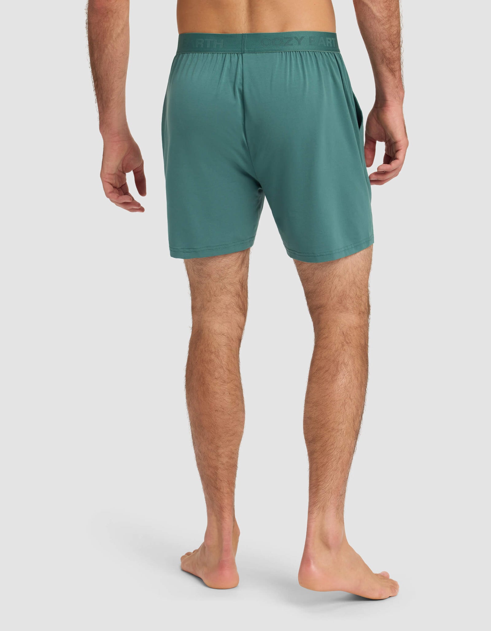 A man stands barefoot on a white background, seen from the back, wearing Cozy Earth's Men's First Layer Pajama Short in teal, featuring an elastic waistband and side pockets.