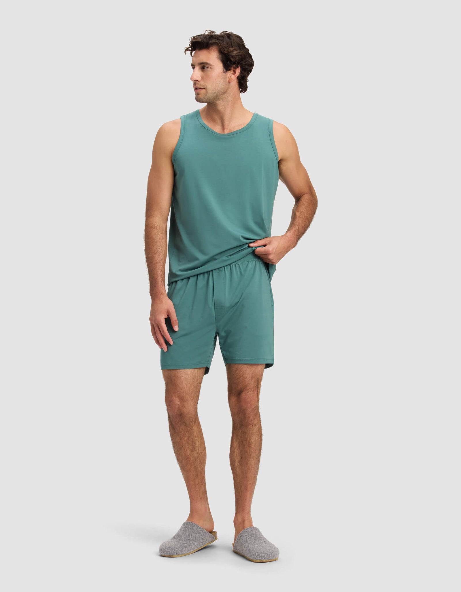A man with short dark hair stands against a plain background wearing Cozy Earth's Men's First Layer Pajama Short set in teal and gray slippers, looking slightly to his left.