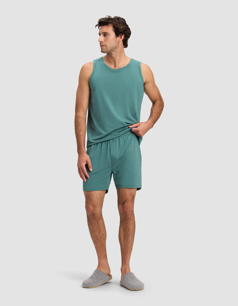 A man with short dark hair stands against a plain background wearing Cozy Earth's Men's First Layer Pajama Short set in teal and gray slippers, looking slightly to his left.