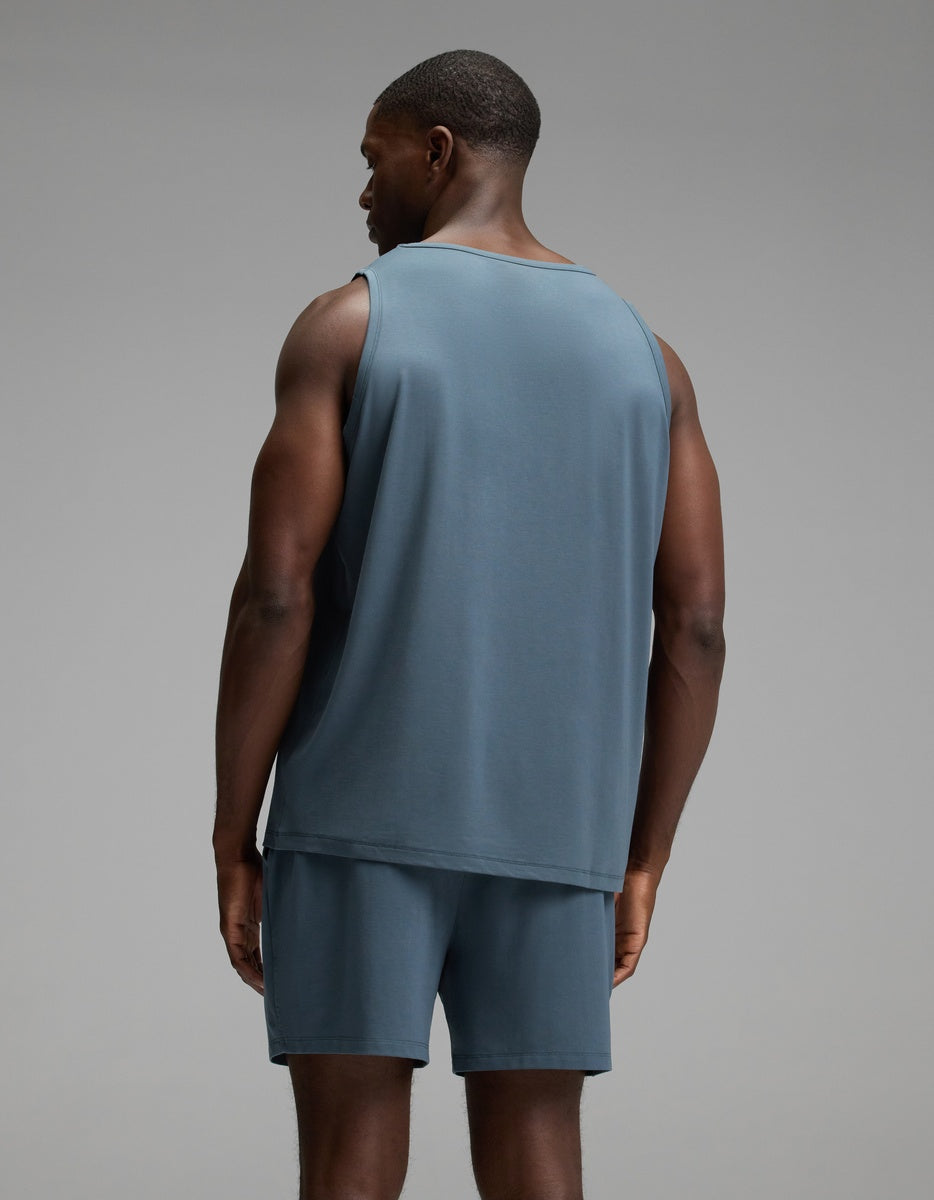 A man stands facing away from the camera, wearing the Cozy Earth Men’s First Layer Pajama Tank in blue, paired with matching blue shorts, against a plain gray background.|Color:Dawn