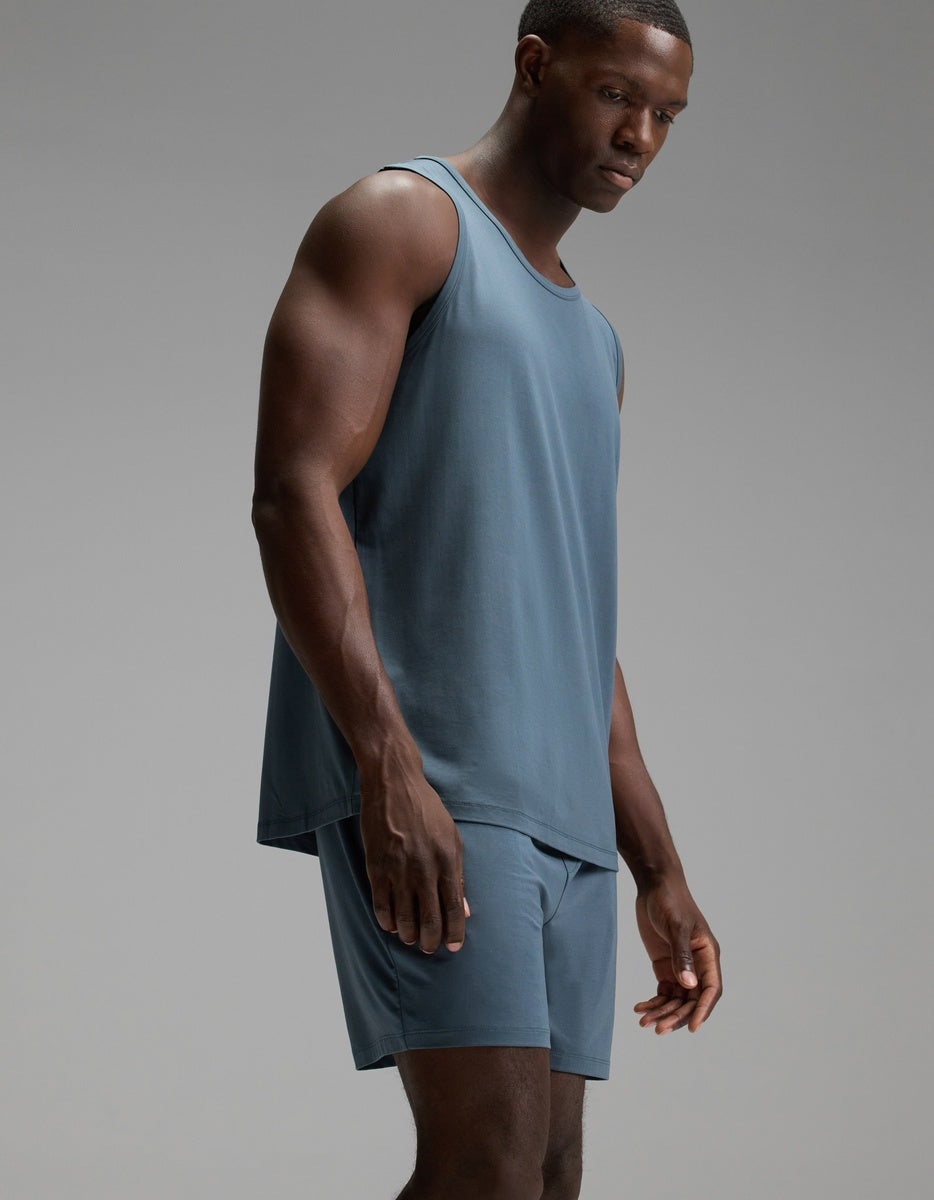 A man stands in a studio wearing the Cozy Earth Men’s First Layer Pajama Tank and matching shorts in blue, looking downward with a neutral expression against a plain gray background.