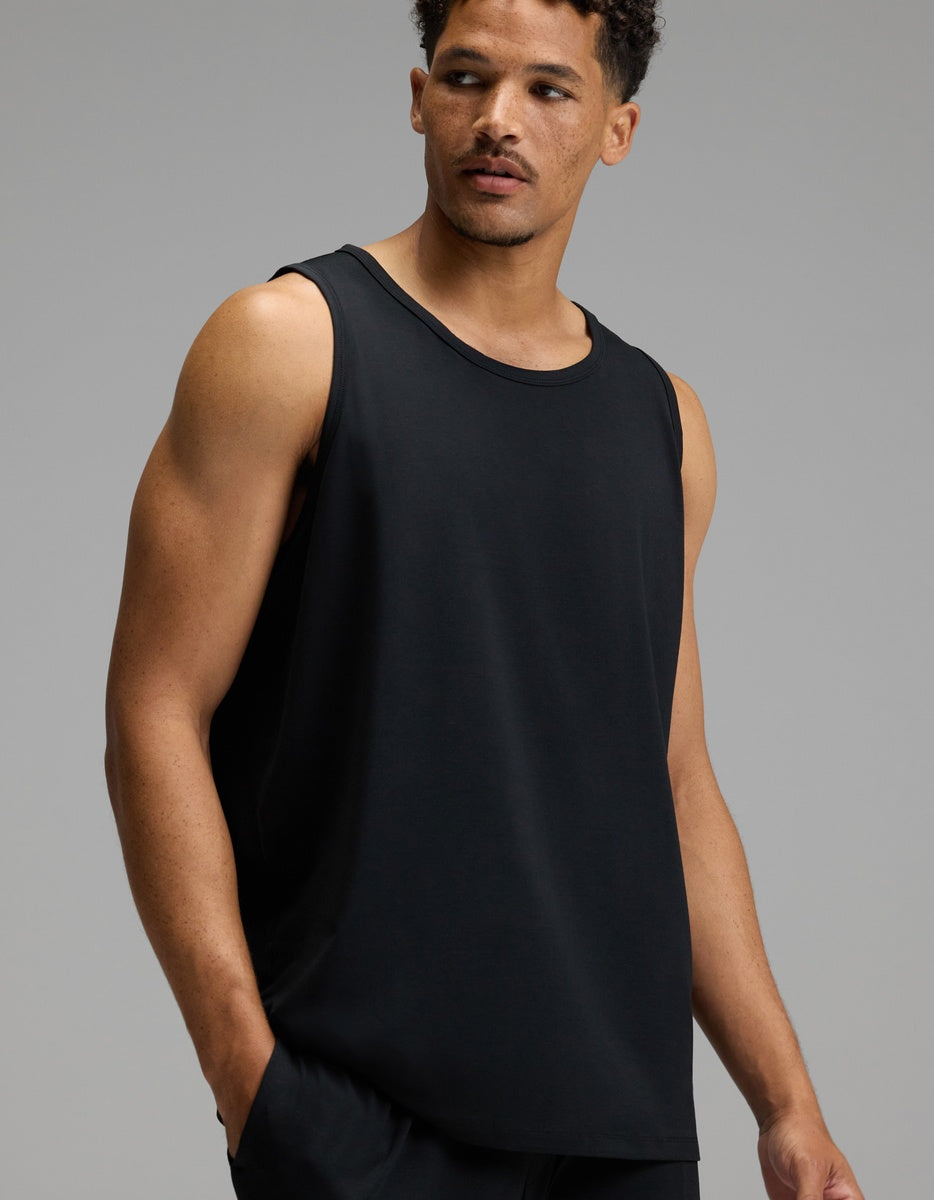 A man wears the Cozy Earth Men’s First Layer Pajama Tank in Jet Black, paired with matching black pants, as he stands against a plain gray background and looks to his left with a neutral expression.|Color:Jet Black