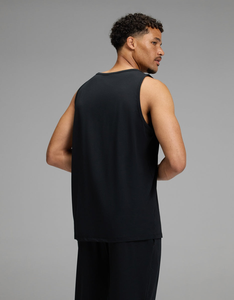 A man with short curly hair is wearing the Cozy Earth Men’s First Layer Pajama Tank in black and matching black pants, facing away from the camera against a plain grey background.