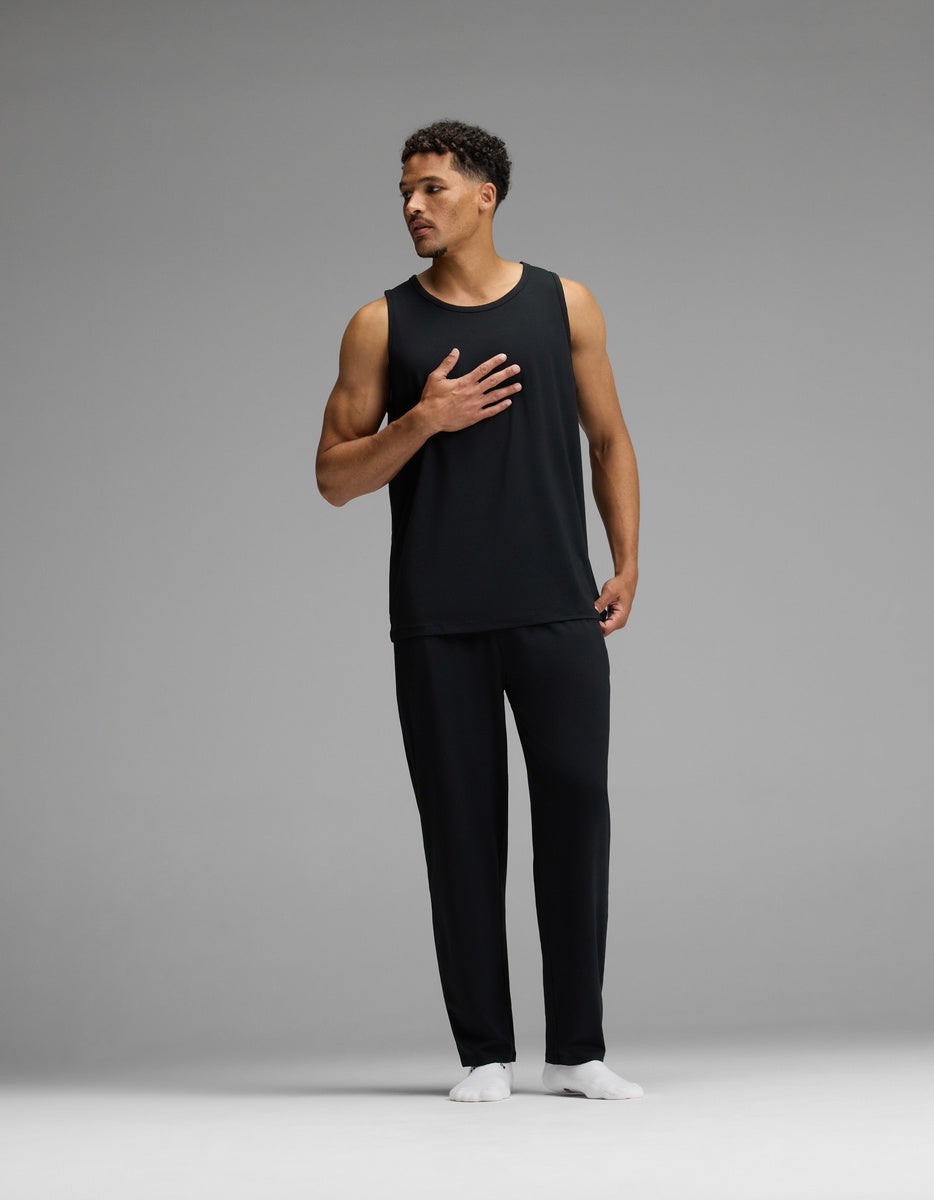 A man wearing the Cozy Earth Men’s First Layer Pajama Tank in black and matching black pants stands with one hand on his chest against a plain gray background, paired with white socks.|Color:Jet Black