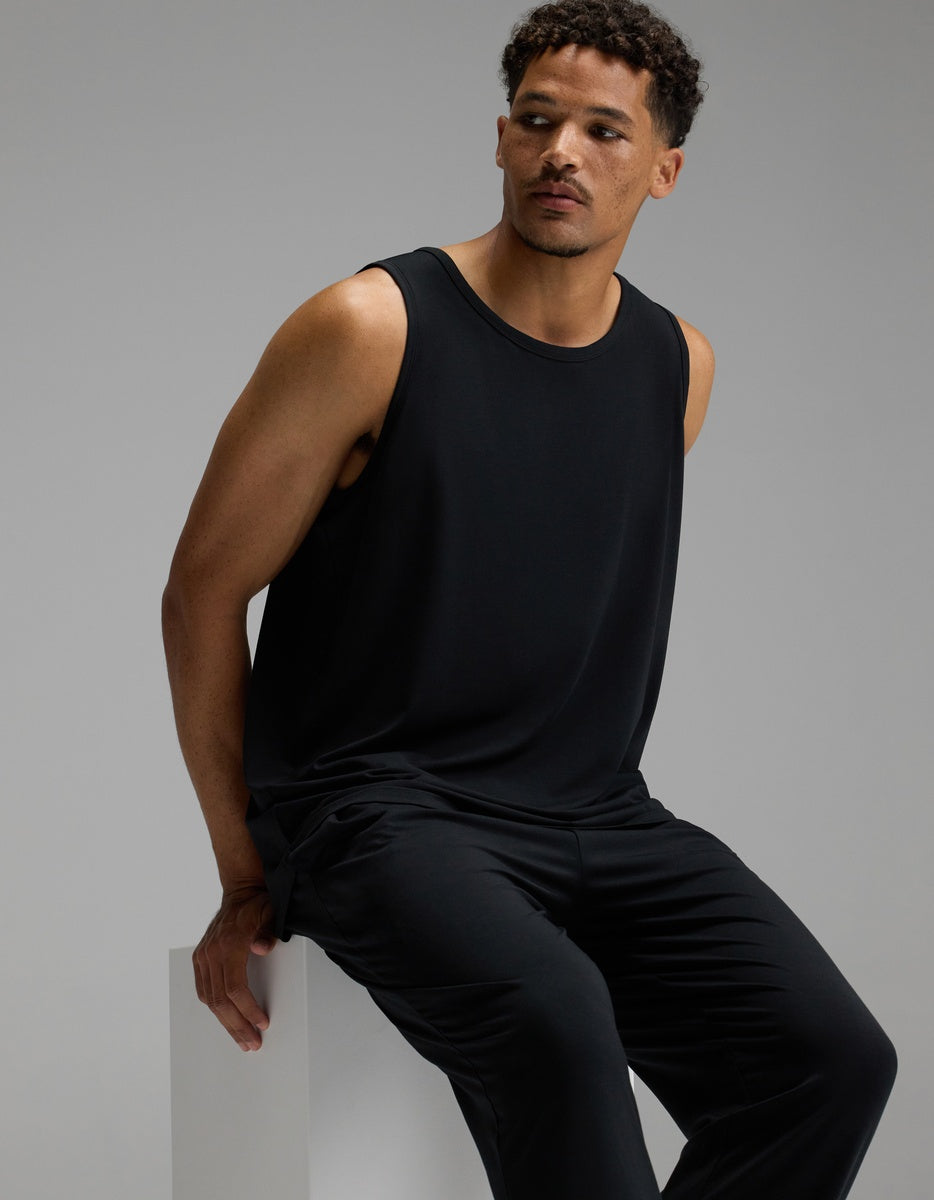 A man wearing the Cozy Earth Men’s First Layer Pajama Tank in black and matching black pants sits on a white block, gazing to the side against a plain gray background.|Color:Jet Black