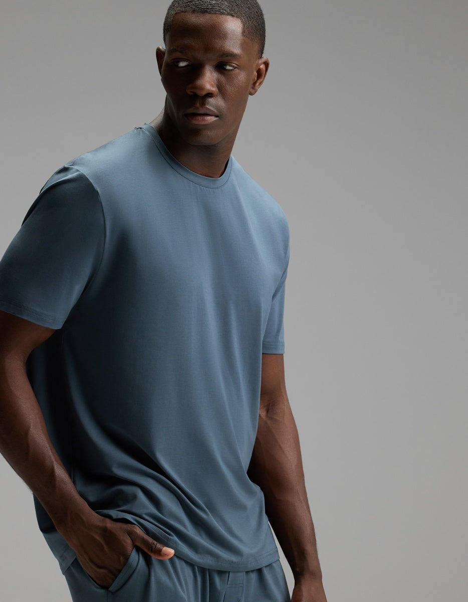 A man wearing the Cozy Earth Men’s First Layer Pajama Short Sleeve Tee in Dawn, along with matching pants, stands with one hand in his pocket and looks to his left against a plain gray background.|Color:Dawn