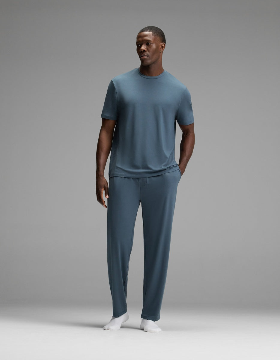 A man stands against a gray background, wearing the Cozy Earth Men’s First Layer Pajama Short Sleeve Tee in blue-gray, paired with matching pants and white socks. His right hand rests at his side while his left hand is tucked into his pocket.