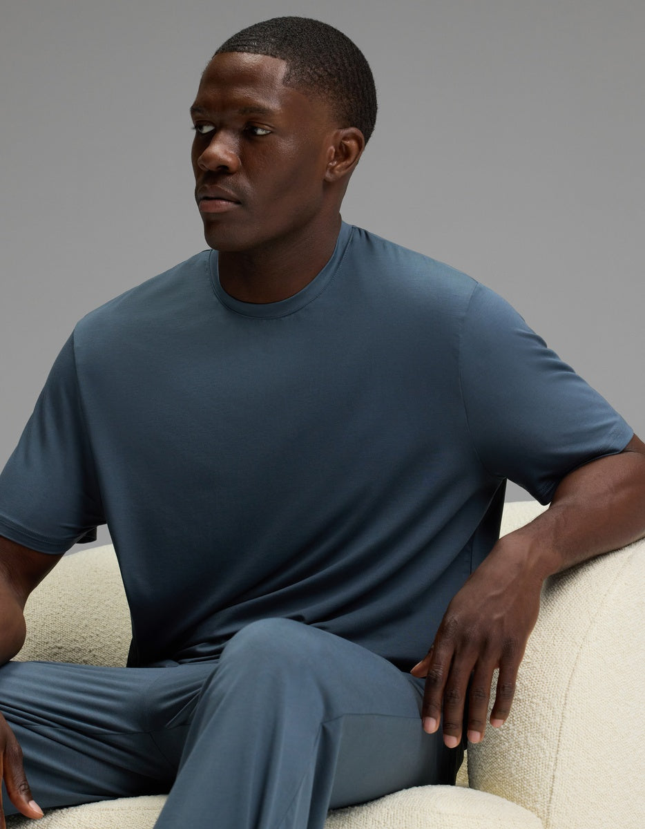 A man wearing the Cozy Earth Men’s First Layer Pajama Short Sleeve Tee sits on a light-colored chair, looking slightly to his left with a neutral expression. The background is plain and gray.