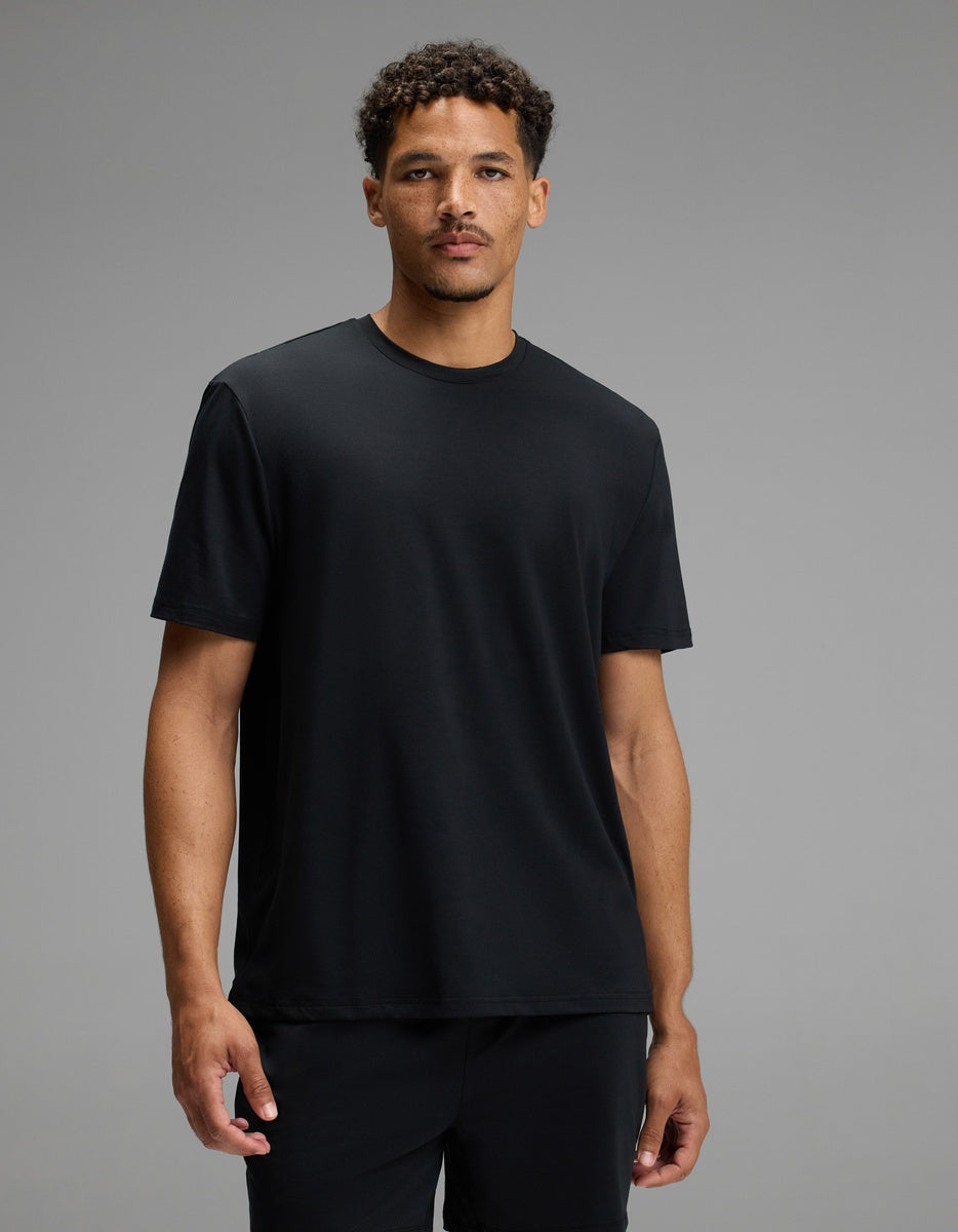 A man with short curly hair wears the Cozy Earth Men’s First Layer Pajama Short Sleeve Tee in Jet Black along with black pants, standing against a plain gray background. He faces forward with a neutral expression.|Color:Jet Black