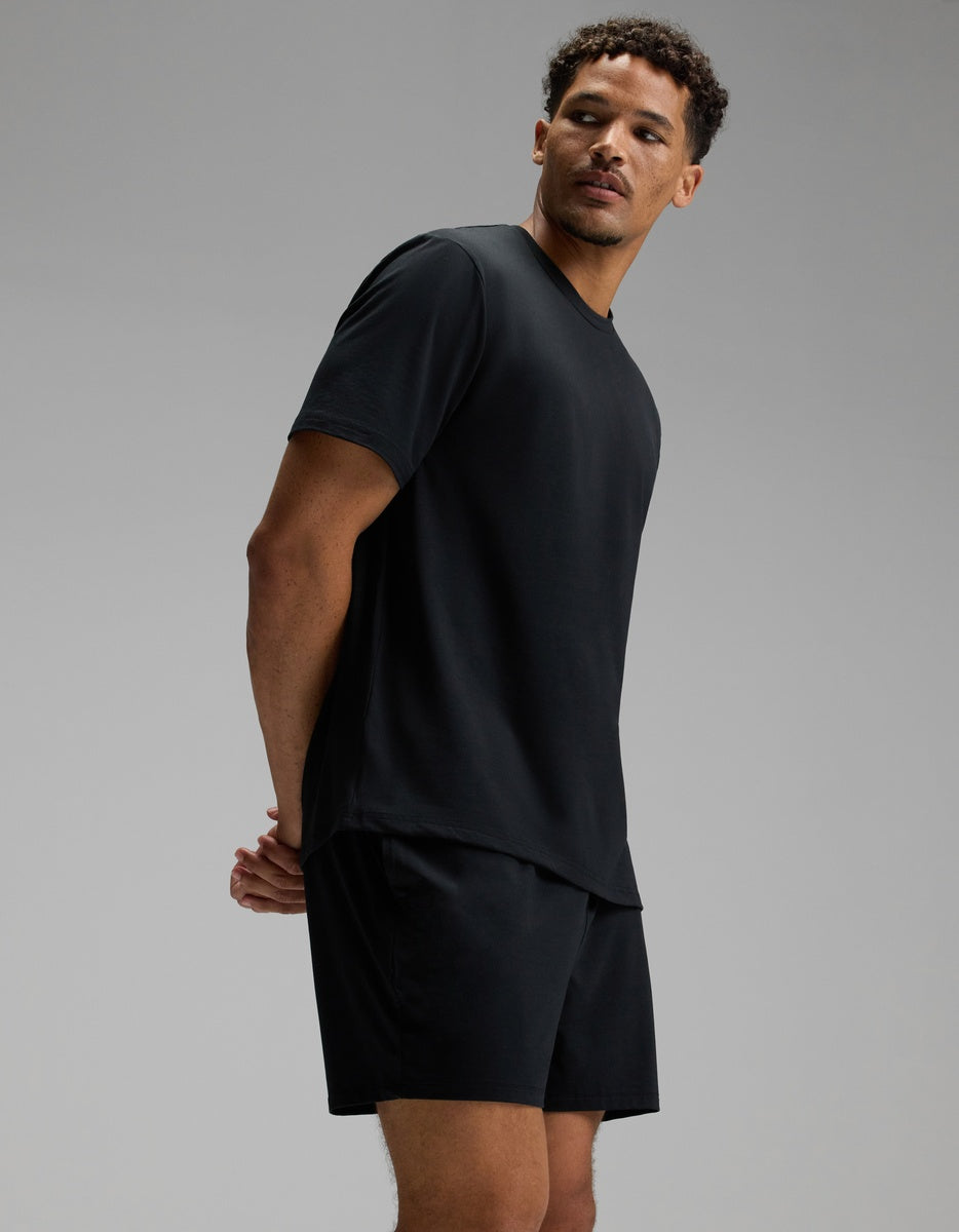 A man wearing a Cozy Earth Men’s First Layer Pajama Short Sleeve Tee and black shorts stands against a plain gray background, looking slightly to the side with his hands behind his back.|Color:Jet Black