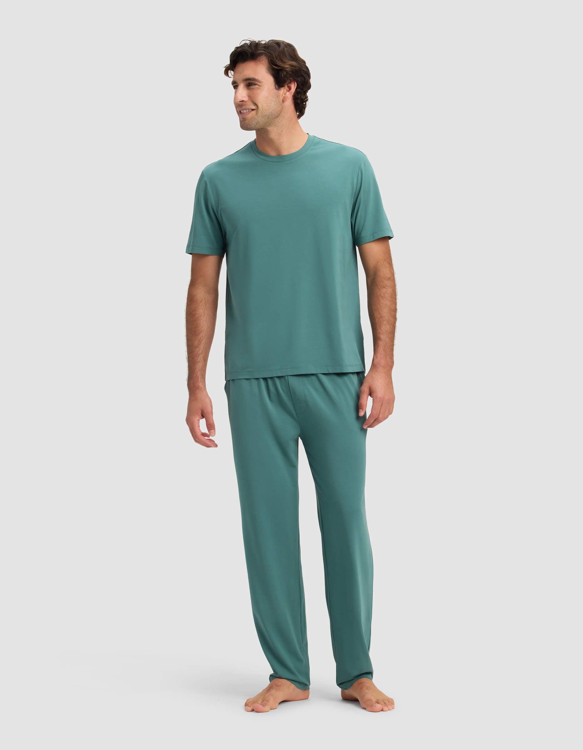 A man smiles while standing barefoot, slightly turned to the side, wearing Cozy Earth's Men's First Layer Pajama Short Sleeve Tee & Pant Set in teal against a plain light background.