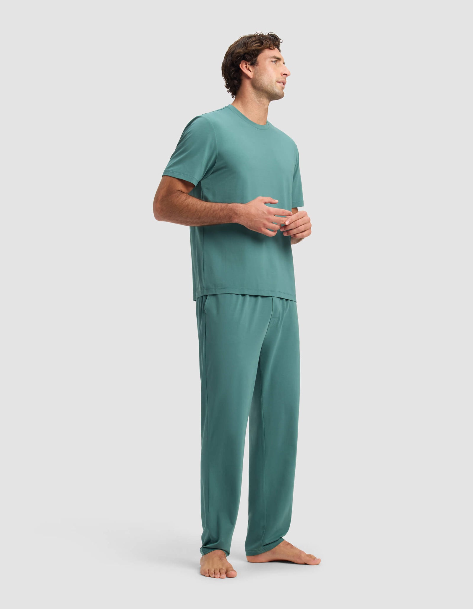 A man stands barefoot on a light background, wearing the Cozy Earth Men's First Layer Pajama Short Sleeve Tee & Pant Set in teal. He looks to the side with his hands loosely clasped.