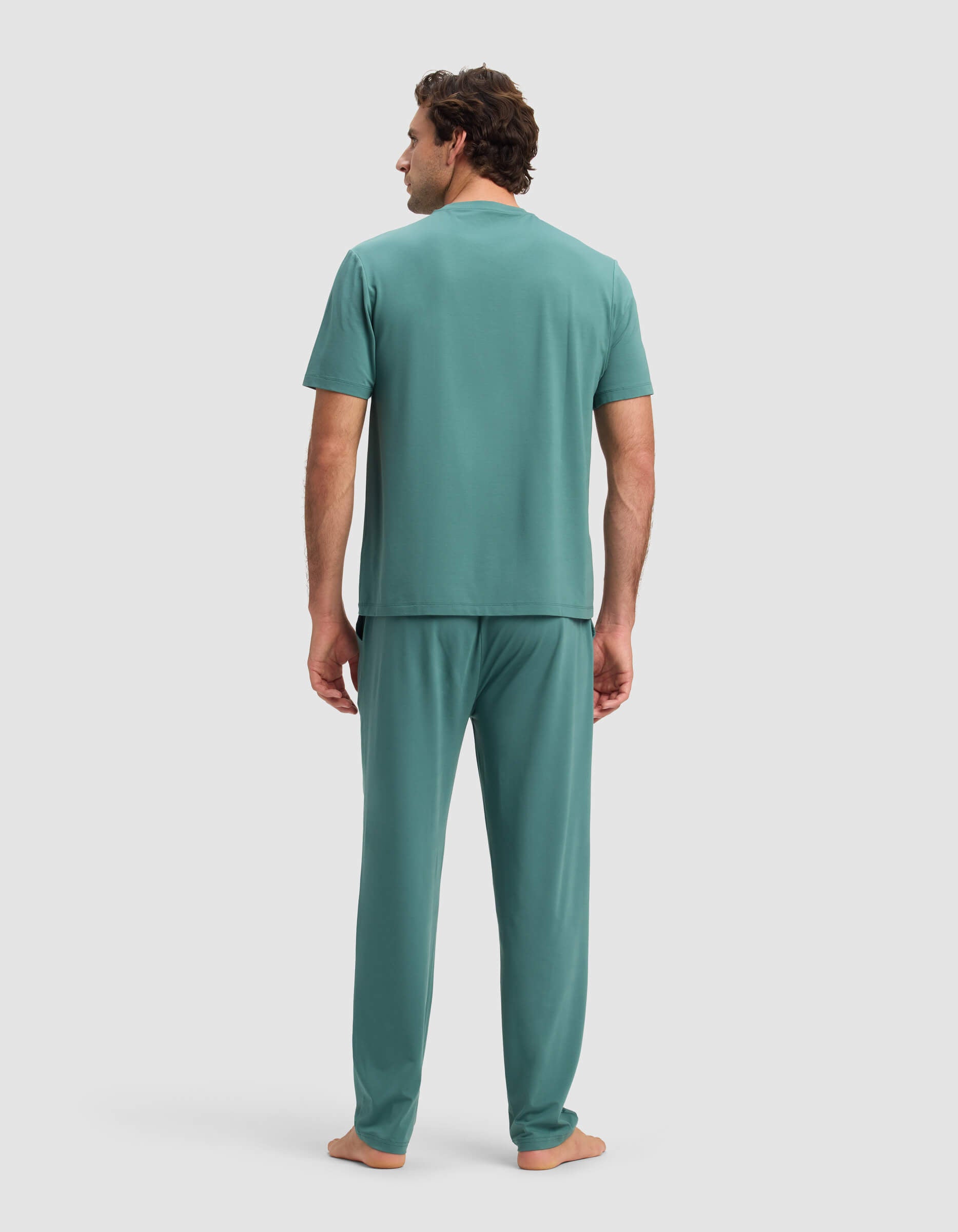 A man stands barefoot facing away, wearing the Cozy Earth Men's First Layer Pajama Short Sleeve Tee & Pant Set in teal, against a plain light gray background.