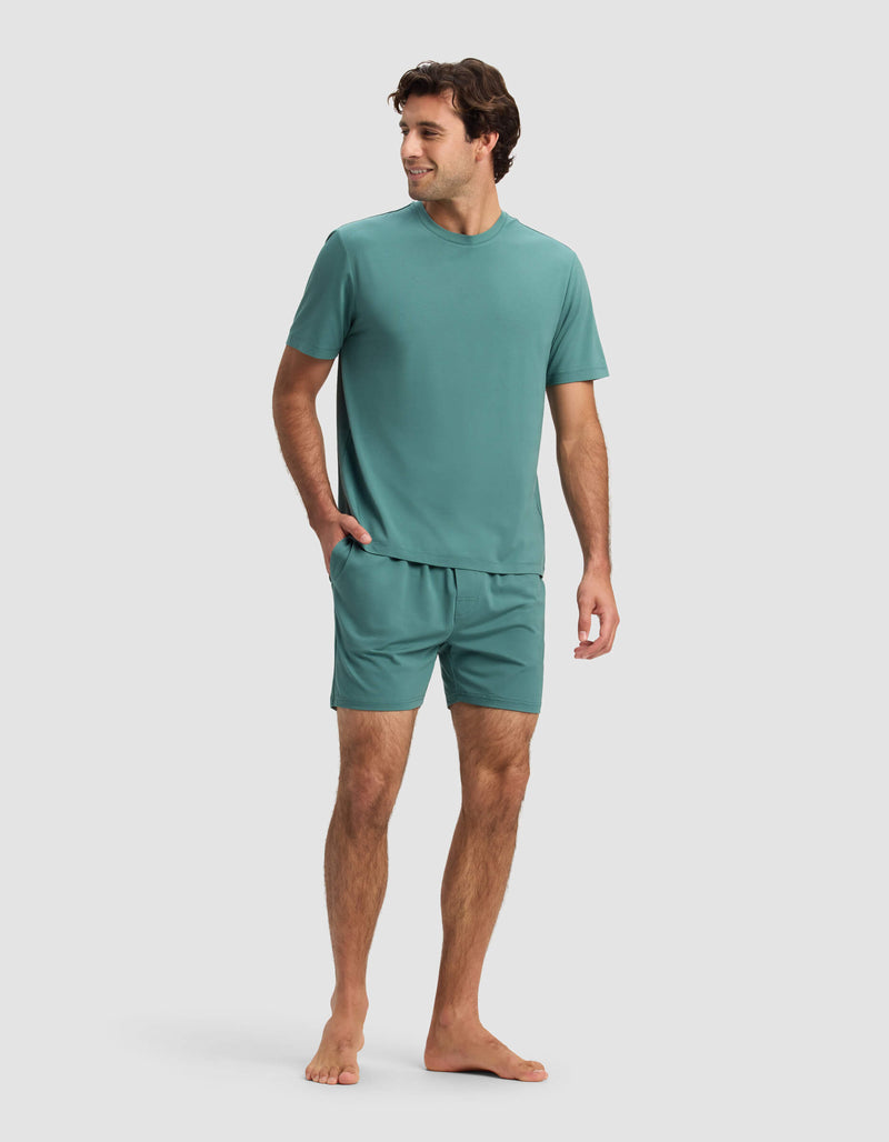 A man stands barefoot against a plain background, wearing the Cozy Earth Men's First Layer Pajama Short Sleeve Tee & Short Set in teal. Facing slightly sideways with one hand in his shorts pocket, he looks relaxed and casual.
