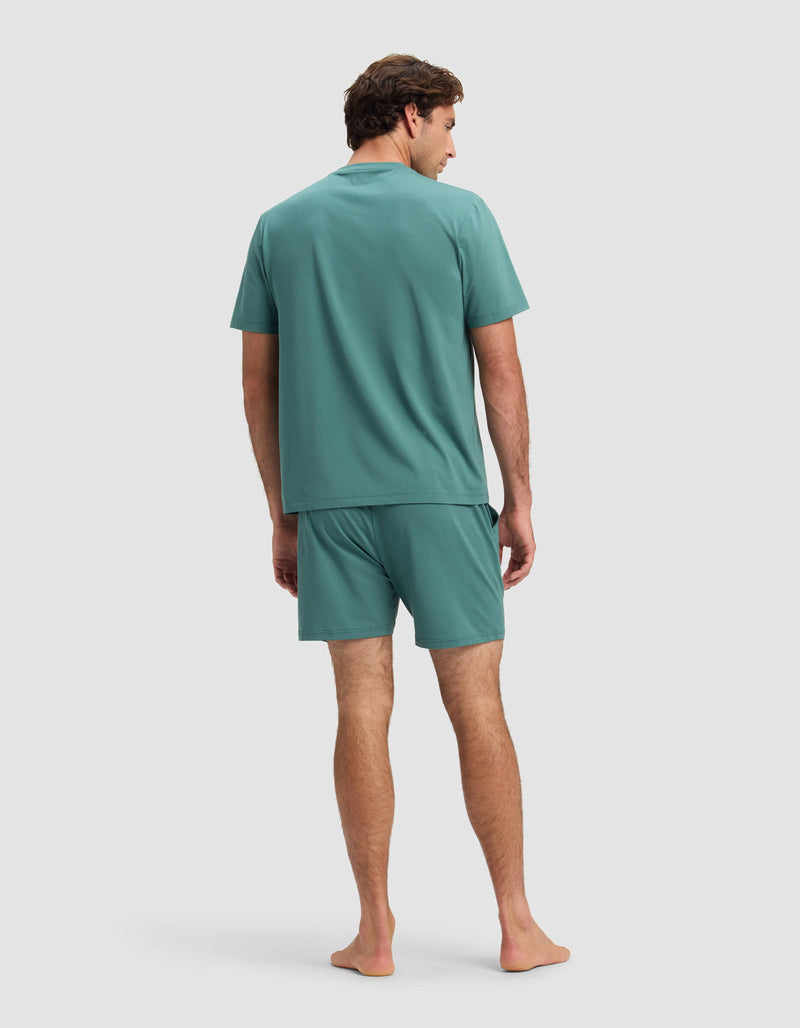 A man stands barefoot facing away, wearing the Cozy Earth Men's First Layer Pajama Short Sleeve Tee & Short Set in teal, against a plain light gray background.