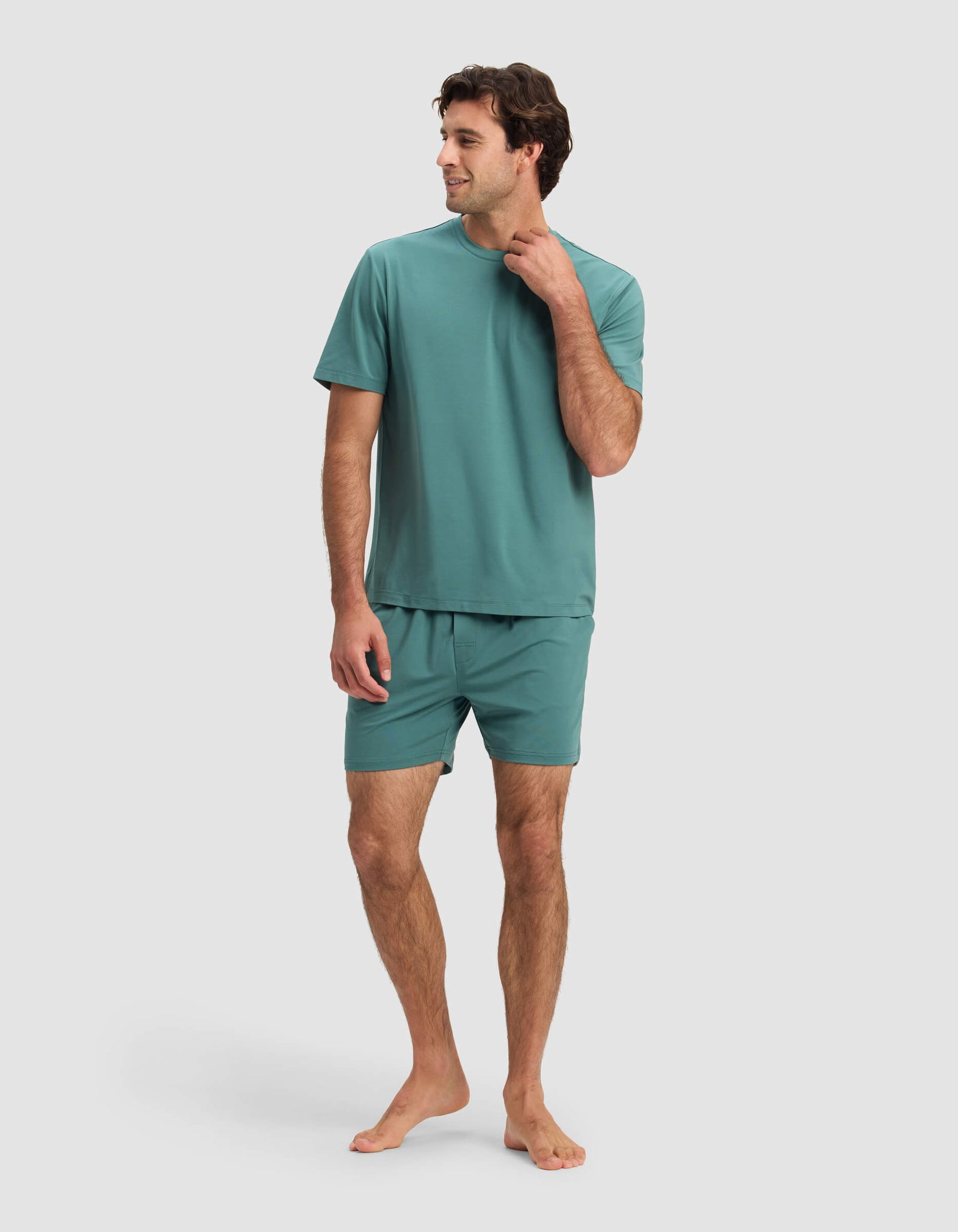 A man stands barefoot against a white background, wearing the Cozy Earth Men's First Layer Pajama Short Sleeve Tee & Short Set in teal, with one hand touching his neck and the other at his side.