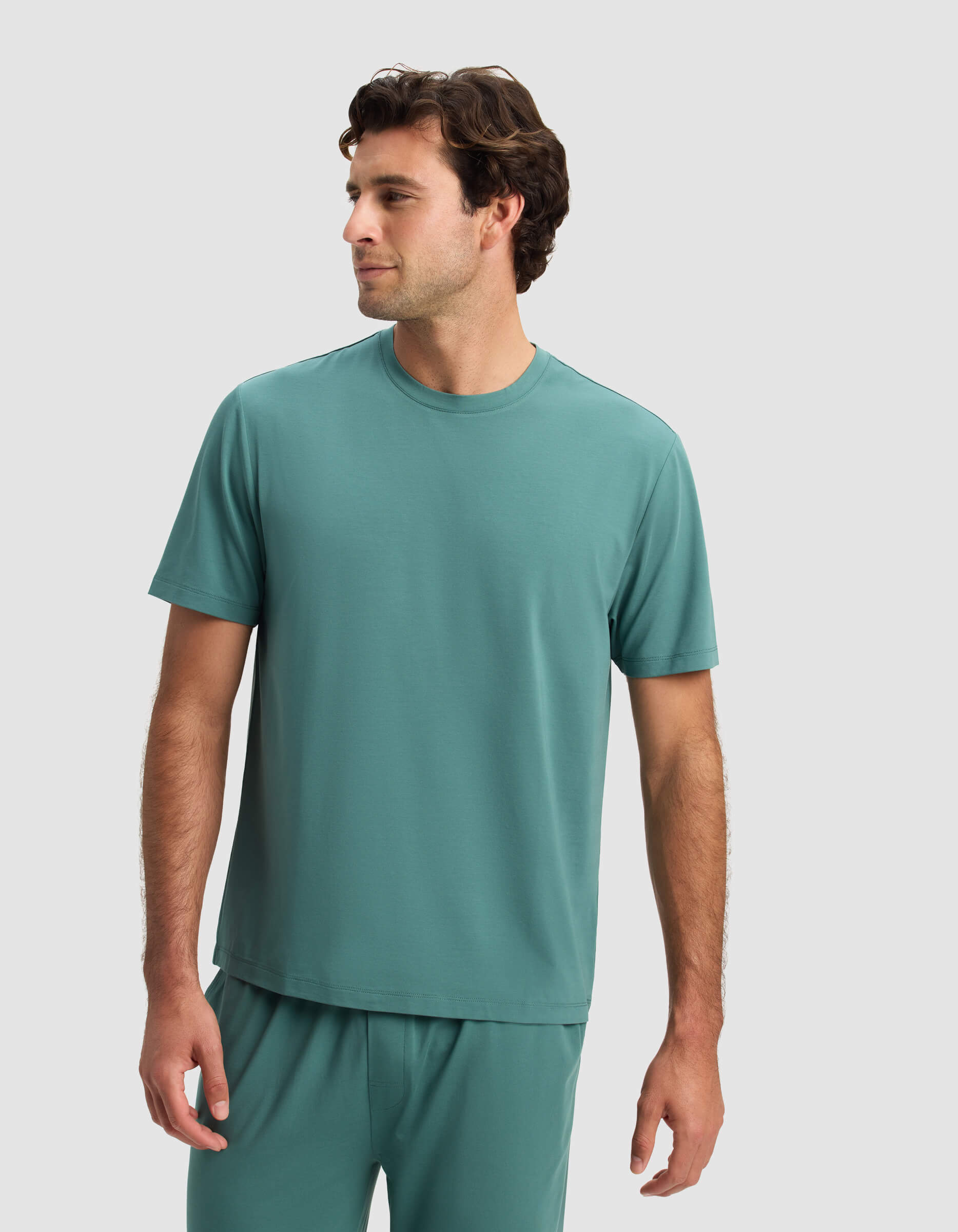 A man wears the Cozy Earth Men's First Layer Pajama Short Sleeve Tee in teal, paired with matching pants, standing against a plain light gray background and looking to his left with a relaxed expression.