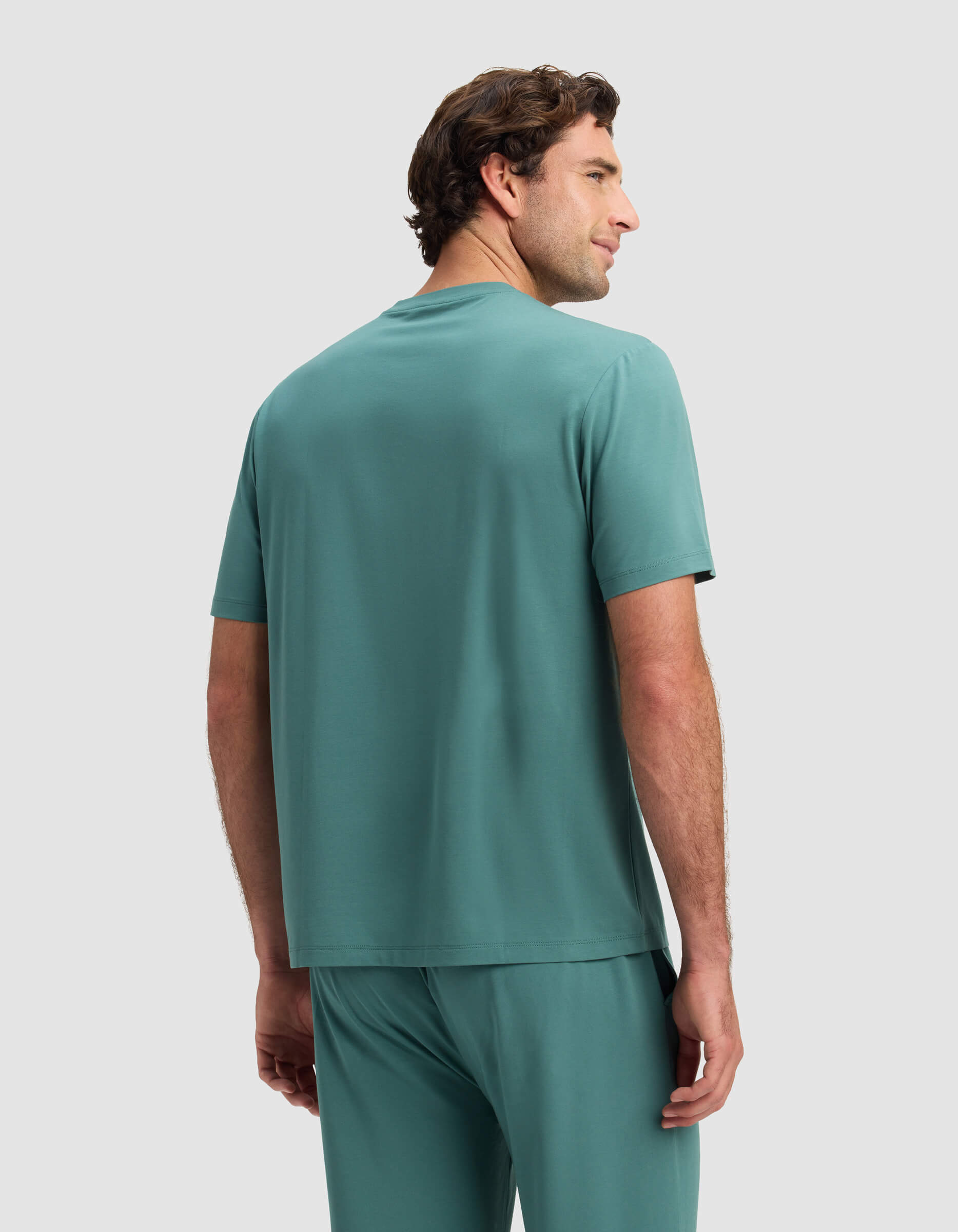 A man stands facing away, wearing a teal Men's First Layer Pajama Short Sleeve Tee by Cozy Earth with matching pants against a plain light gray background.