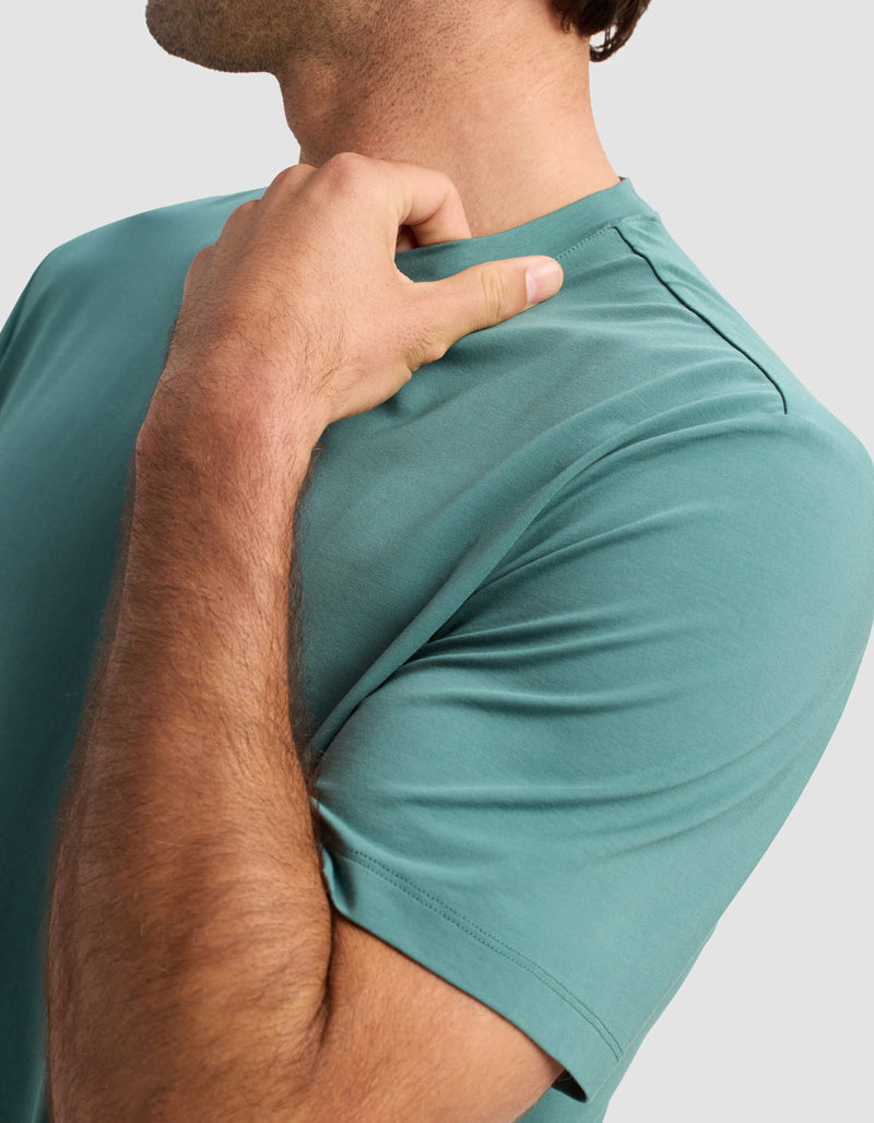 A man is shown from the shoulders up wearing the Cozy Earth Men's First Layer Pajama Short Sleeve Tee in teal. He touches the shirt's collar with his right hand against a simple light background.