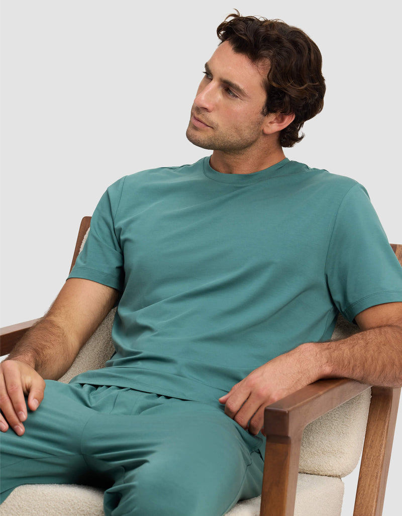 A man with wavy brown hair sits in a cushioned wooden chair, wearing the Cozy Earth Men's First Layer Pajama Short Sleeve Tee and matching pants. He looks relaxed, gazing to the side against a plain light gray background.