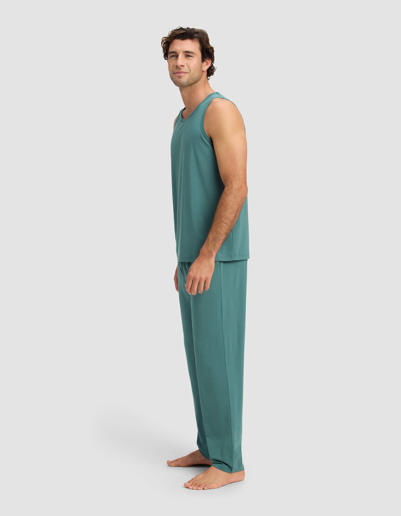 A man stands barefoot on a plain background, wearing the Cozy Earth Men's First Layer Pajama Tank & Pant Set in teal, looking at the camera with a relaxed expression.
