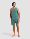 A man stands barefoot in Cozy Earth's Men's First Layer Pajama Tank & Short Set in teal, one hand in his pocket, smiling and looking to the side against a plain light gray background.|Color:Sea Green