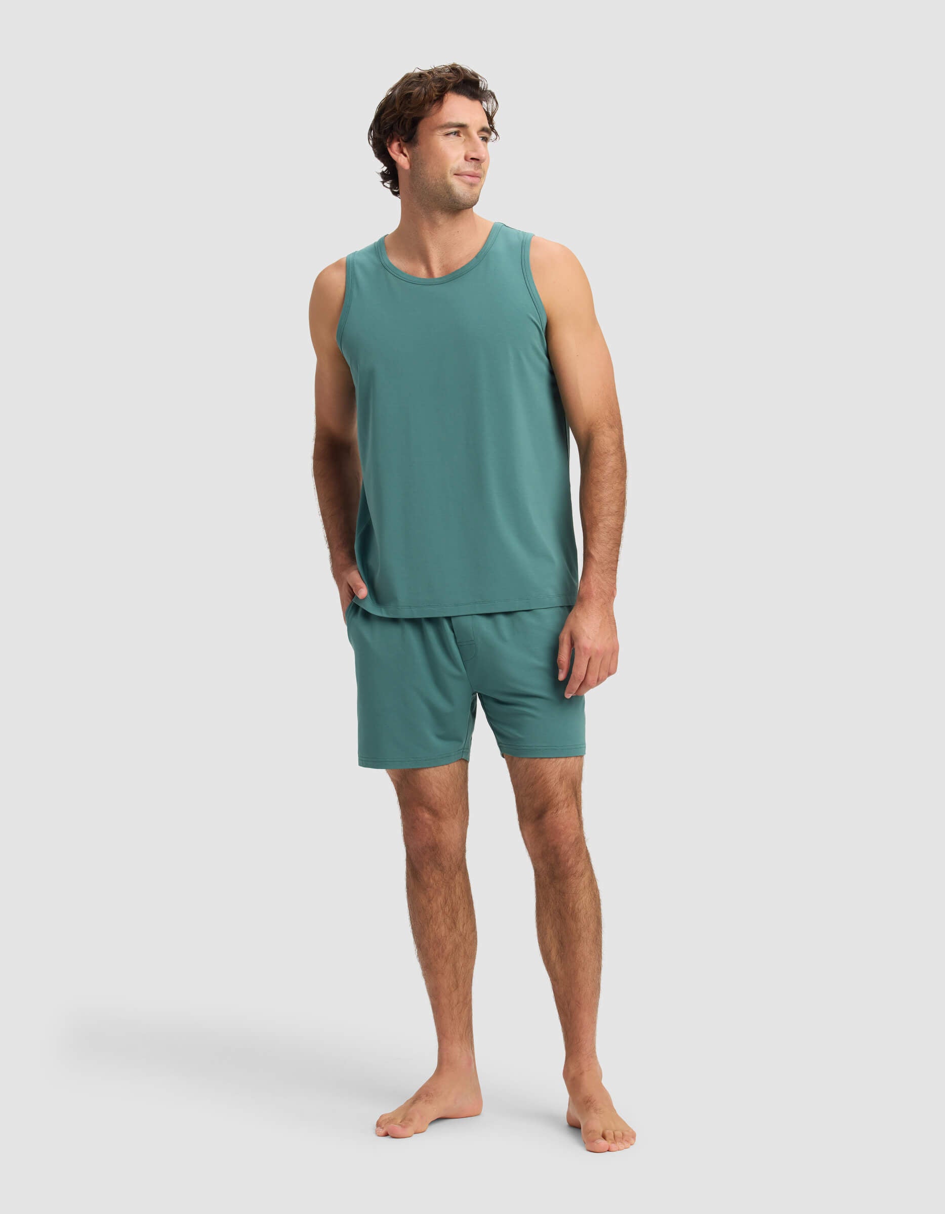 A man stands barefoot in Cozy Earth's Men's First Layer Pajama Tank & Short Set in teal, one hand in his pocket, smiling and looking to the side against a plain light gray background.|Color:Sea Green