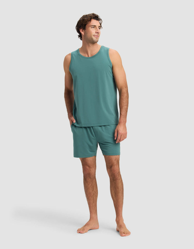 A man stands barefoot in Cozy Earth's Men's First Layer Pajama Tank & Short Set in teal, one hand in his pocket, smiling and looking to the side against a plain light gray background.