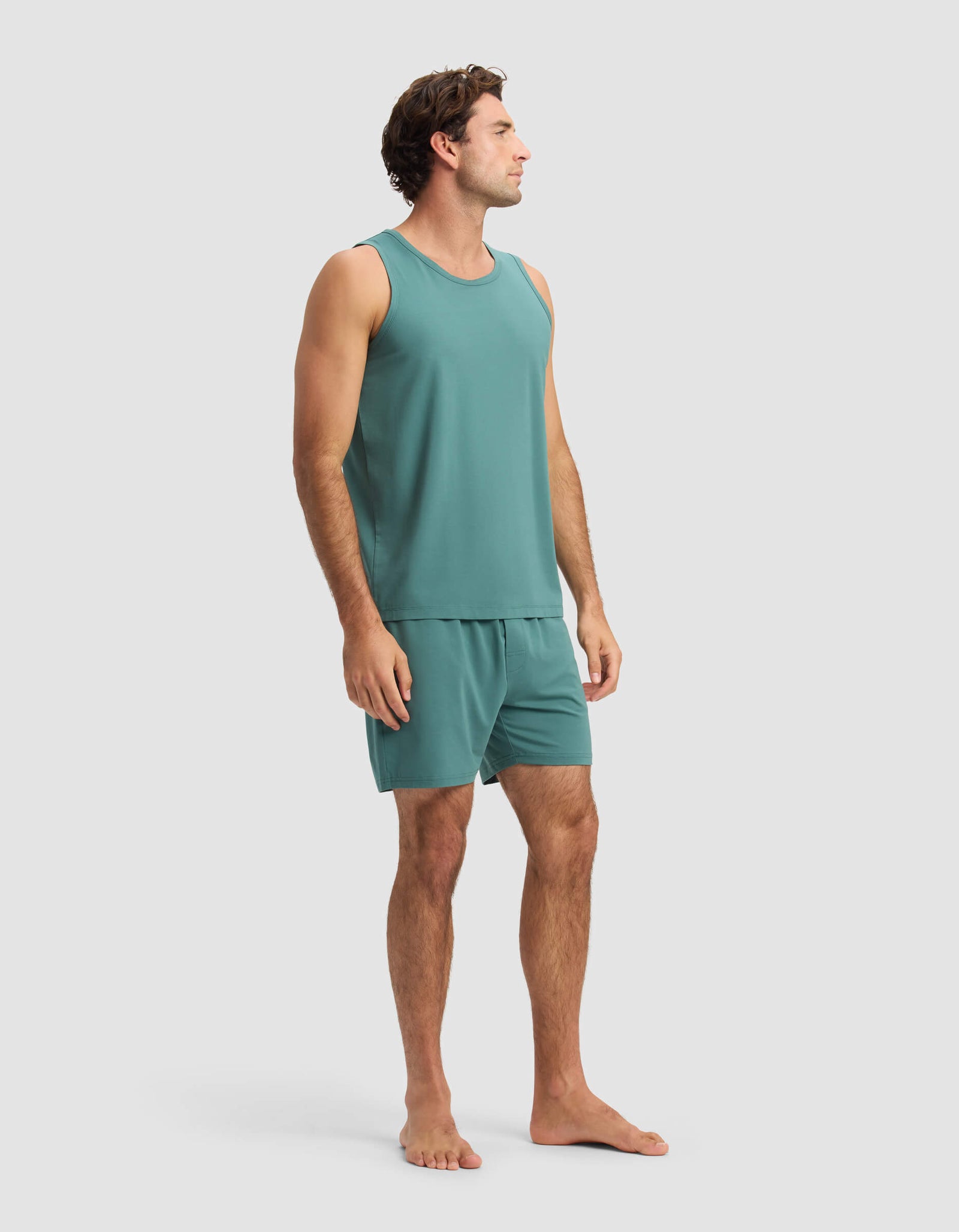A man stands barefoot in the Cozy Earth Men's First Layer Pajama Tank & Short Set, facing slightly to the side against a plain, light background.