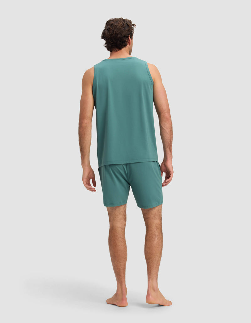 A man with short, dark hair stands barefoot, facing away from the camera, wearing the Men's First Layer Pajama Tank & Short Set by Cozy Earth in teal against a plain light gray background.