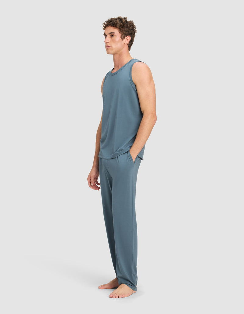 A man stands barefoot against a light gray background, wearing Cozy Earth's Men's First Layer Pajama Tank & Pant Set in blue-gray, with his hands in his pockets.