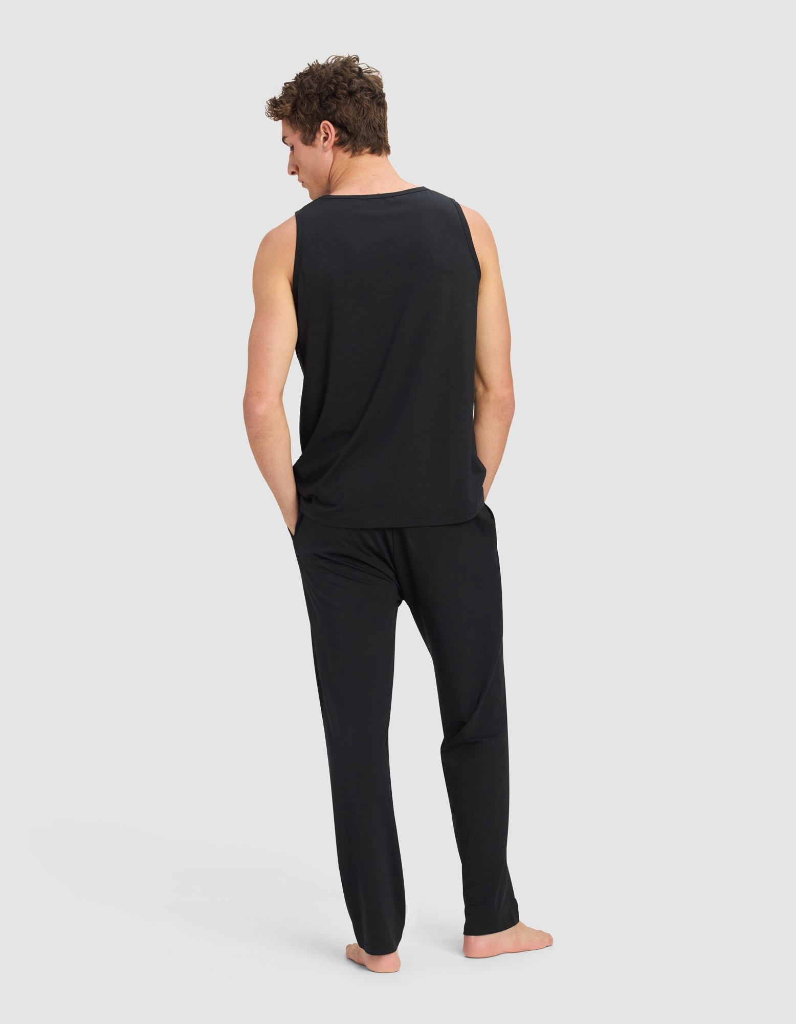 A man stands barefoot, wearing the Cozy Earth Men's First Layer Pajama Tank & Pant Set in black, with his hands in his pockets, facing away from the camera against a plain light gray background.|Color:Jet Black