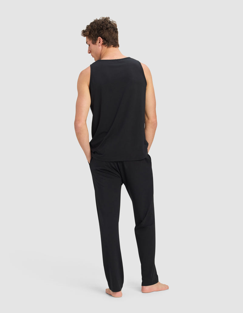 A man stands barefoot, wearing the Cozy Earth Men's First Layer Pajama Tank & Pant Set in black, with his hands in his pockets, facing away from the camera against a plain light gray background.