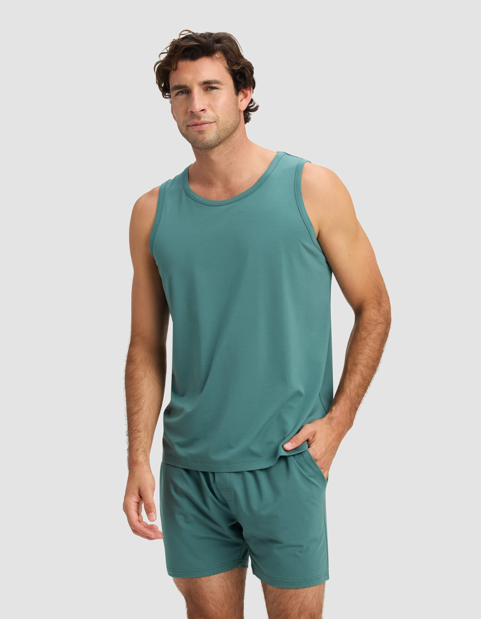 A man with short brown hair models the Cozy Earth Men's First Layer Pajama Tank and matching shorts in teal, standing against a plain light gray background with one hand in his pocket.