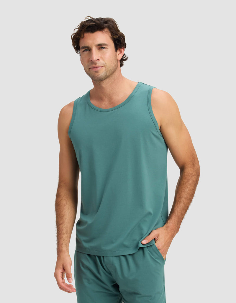 A man stands against a light gray background, smiling slightly with one hand in his pocket, wearing the Cozy Earth Men's First Layer Pajama Tank & Pant Set.