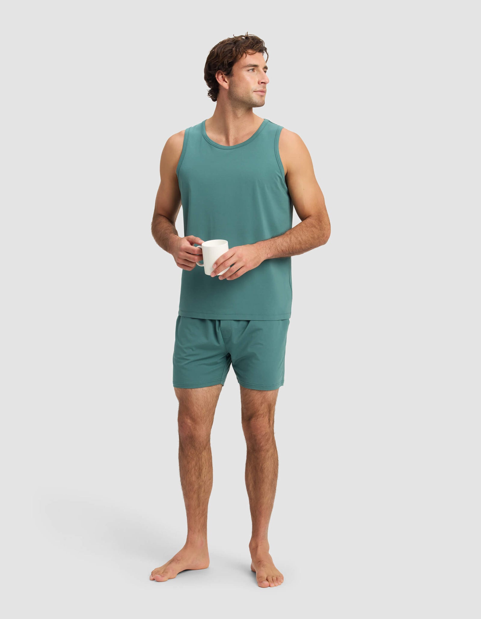 A man wearing the Cozy Earth Men's First Layer Pajama Tank and matching shorts stands barefoot, holding a white mug in both hands, gazing slightly to his left against a plain light background.