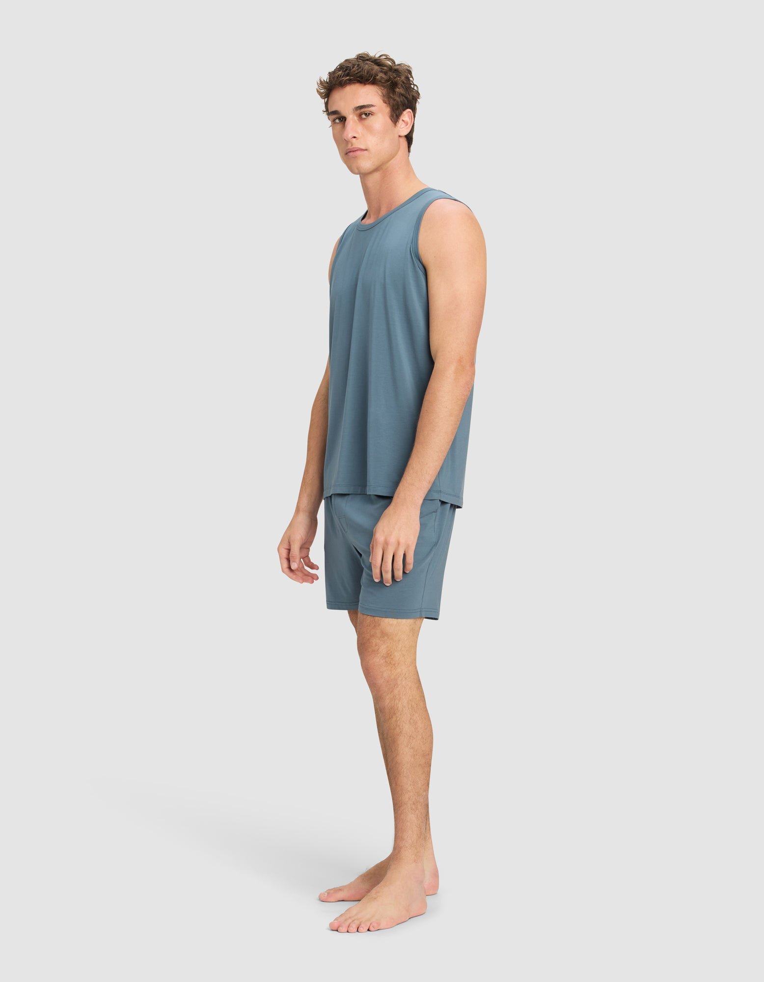 A young man stands barefoot on a neutral background, wearing the Men's First Layer Pajama Tank & Short Set from Cozy Earth in blue. He faces slightly to the side with a relaxed expression and a comfortable, casual look.