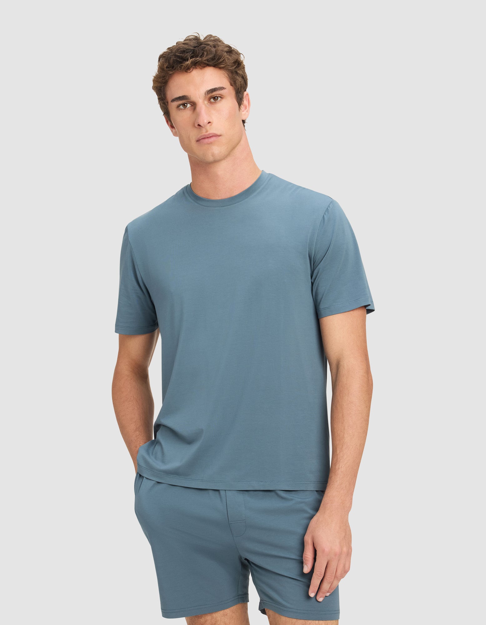 A young man with short curly hair wears the Cozy Earth Men's First Layer Pajama Short Sleeve Tee and matching shorts, standing in front of a plain light gray background with one hand in his pocket.