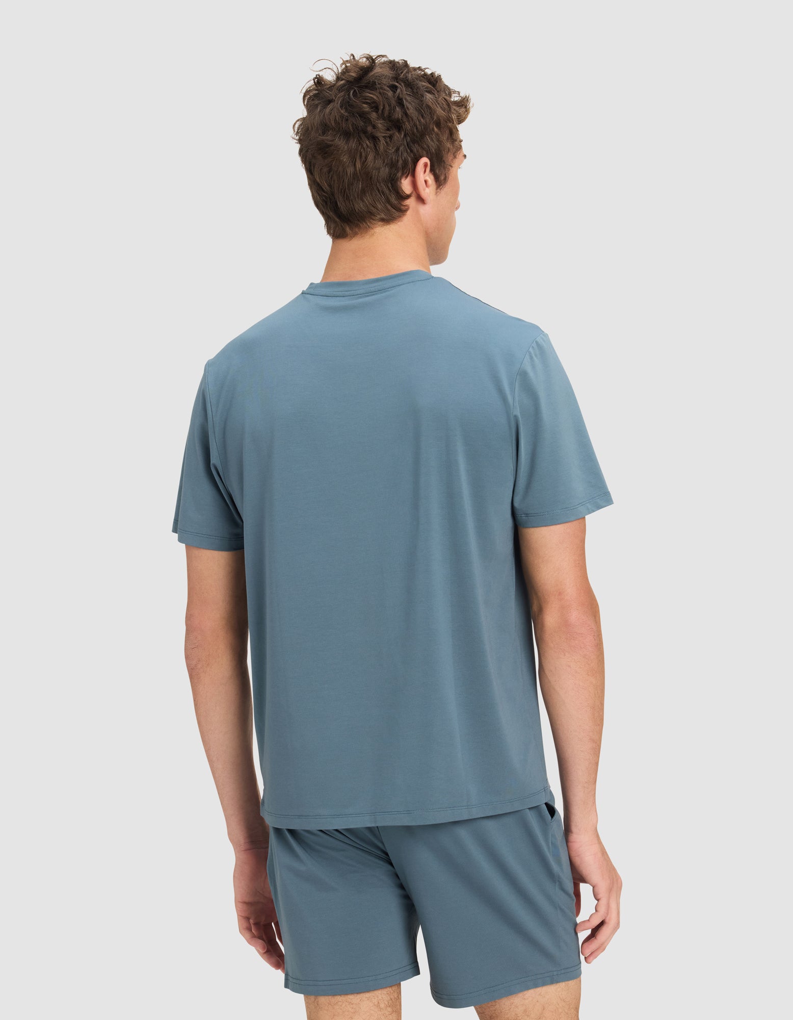 A man with short brown hair, seen from behind, is wearing the Cozy Earth Men's First Layer Pajama Short Sleeve Tee in light blue, paired with matching shorts against a plain light background.