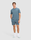 A man stands against a plain background wearing Cozy Earth's Men's First Layer Pajama Short Sleeve Tee in blue, matching blue shorts, and white socks with light blue toes and heels. He has one hand in his pocket and a neutral expression.|Color:Dawn