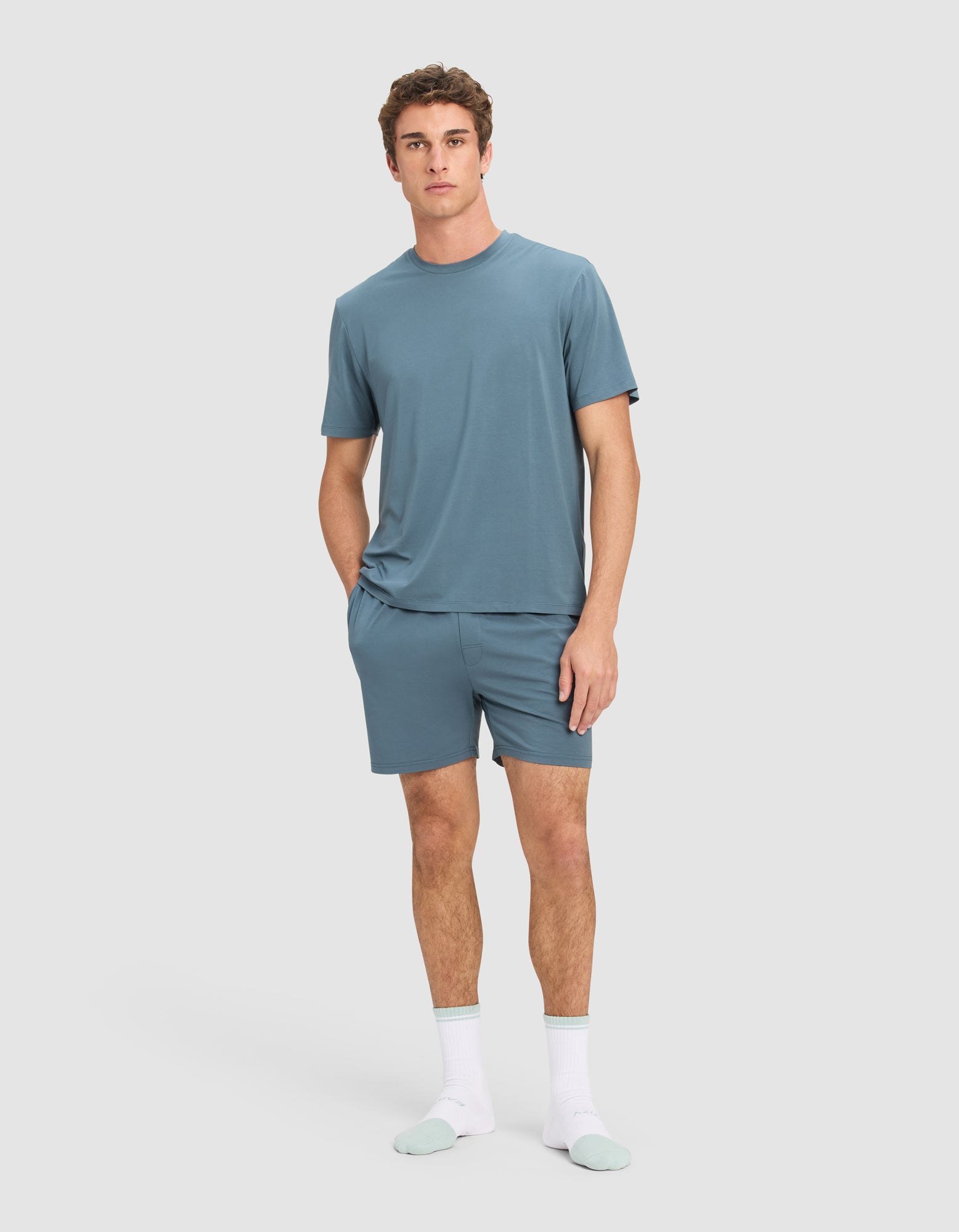 A man stands against a plain background wearing Cozy Earth's Men's First Layer Pajama Short Sleeve Tee in blue, matching blue shorts, and white socks with light blue toes and heels. He has one hand in his pocket and a neutral expression.