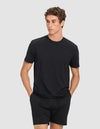 A man wears the Cozy Earth Men's First Layer Pajama Short Sleeve Tee in black, paired with black shorts, standing with hands in his pockets and looking to the side against a light gray background.|Color:Jet Black