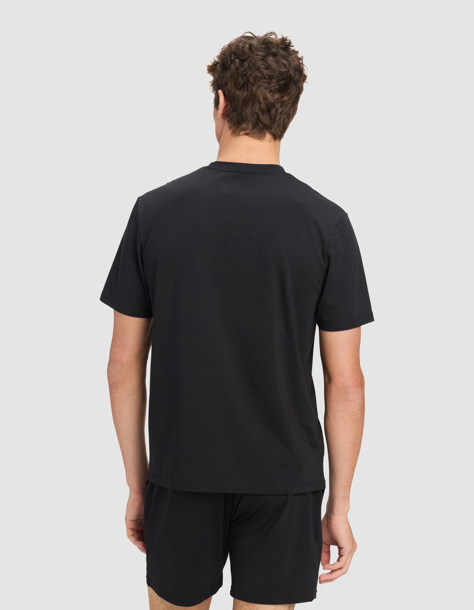 A man with curly brown hair stands against a light gray background, facing away from the camera, wearing a black Men's First Layer Pajama Short Sleeve Tee by Cozy Earth and black shorts.