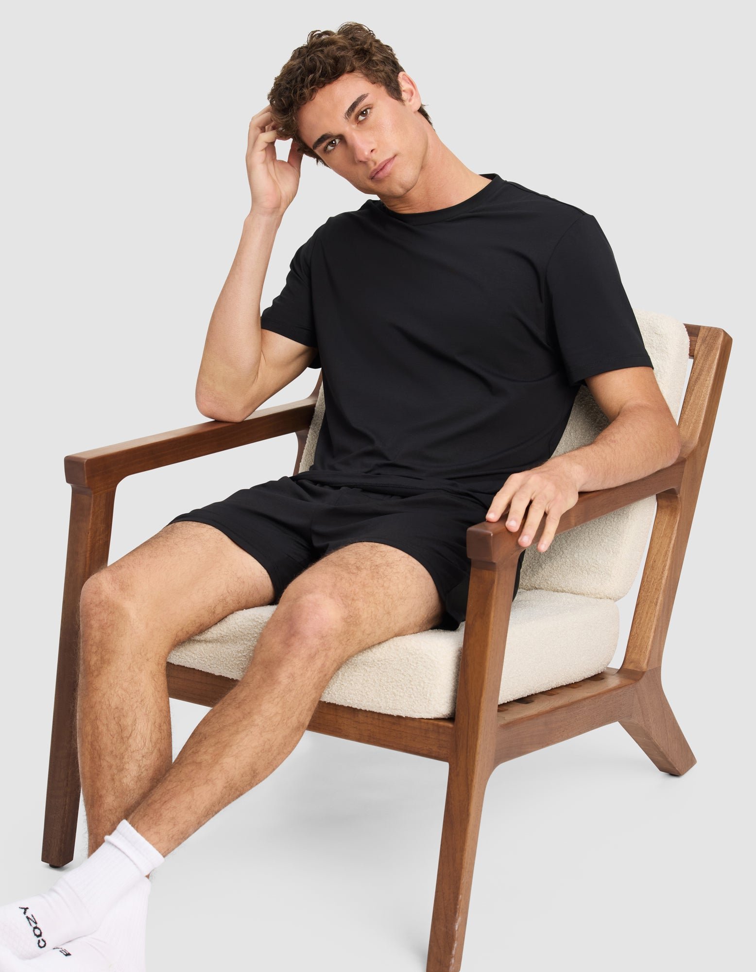 A young man wears the Cozy Earth Men's First Layer Pajama Short Sleeve Tee and shorts as he relaxes in a modern wooden chair with cream cushions, resting one hand on his head and the other on the armrest, paired with white socks.|Color:Jet Black