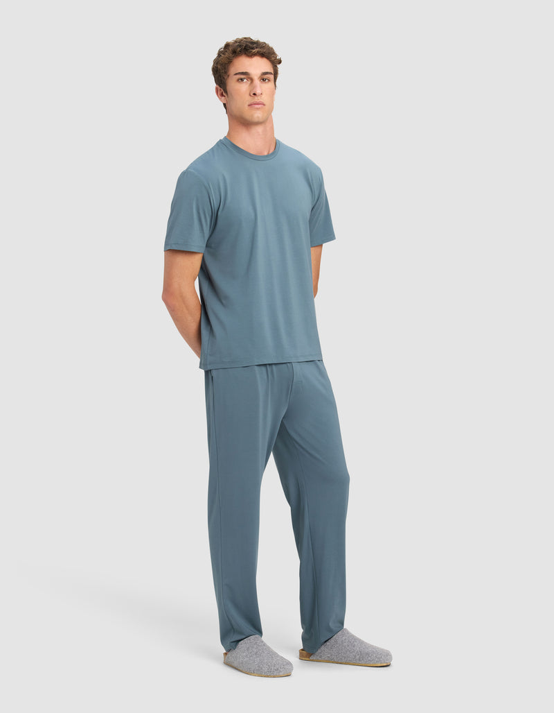 A man with short curly hair stands against a light background wearing the Cozy Earth Men's First Layer Pajama Short Sleeve Tee & Pant Set in blue and gray slippers, with his hands behind his back.